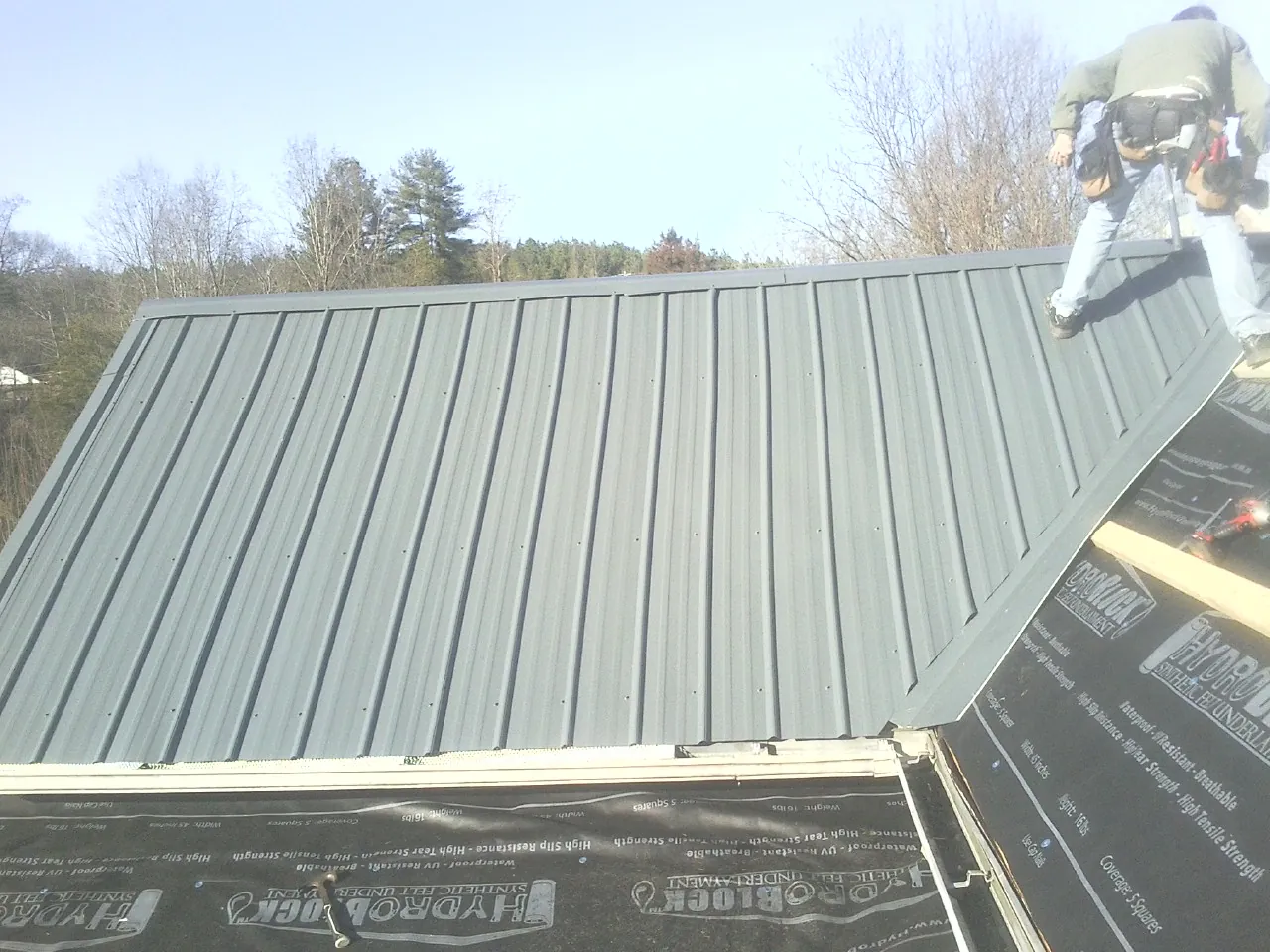 Crew installing dark green standing seam panels over synthetic underlayment in Lilly
