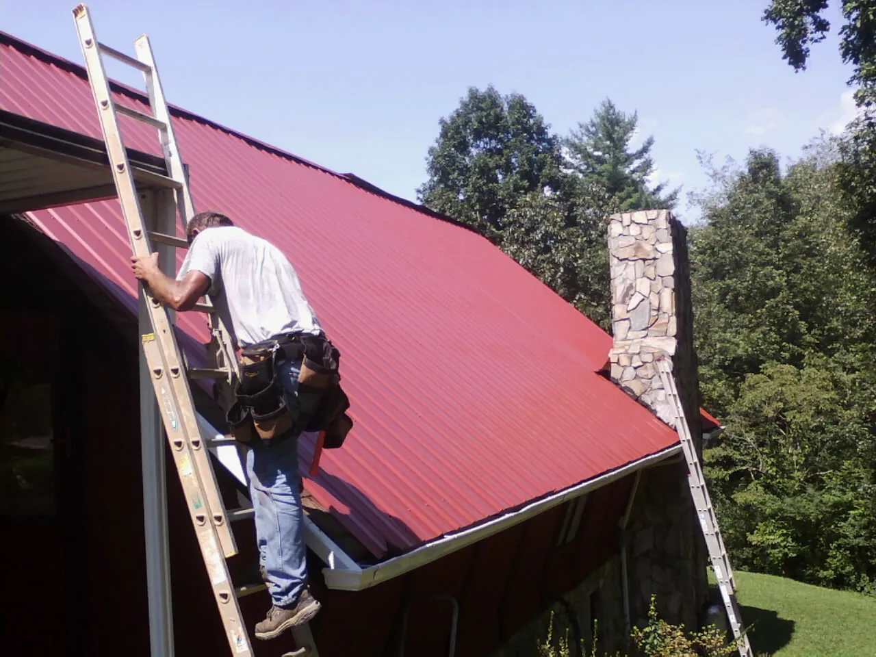 Roofer on a ladder beside a red metal roof and stone chimney — Soffit & Fascia Repair by Roofing Brothers in Lilly